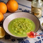 Nage de concombre froide à l'orange et au zaatar dans une assiette rayée rouge et blanc, posée sur une nappe blanche brodée au point de Fès bleu, entourée de trois oranges, un demi citron, une bouteille d'eau de fleur d'oranger et un ramequin de zaatar